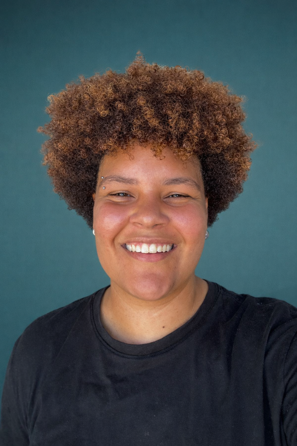 Pessoa sorridente com cabelo encaracolado vestindo uma camisa preta contra um fundo azul-petróleo.