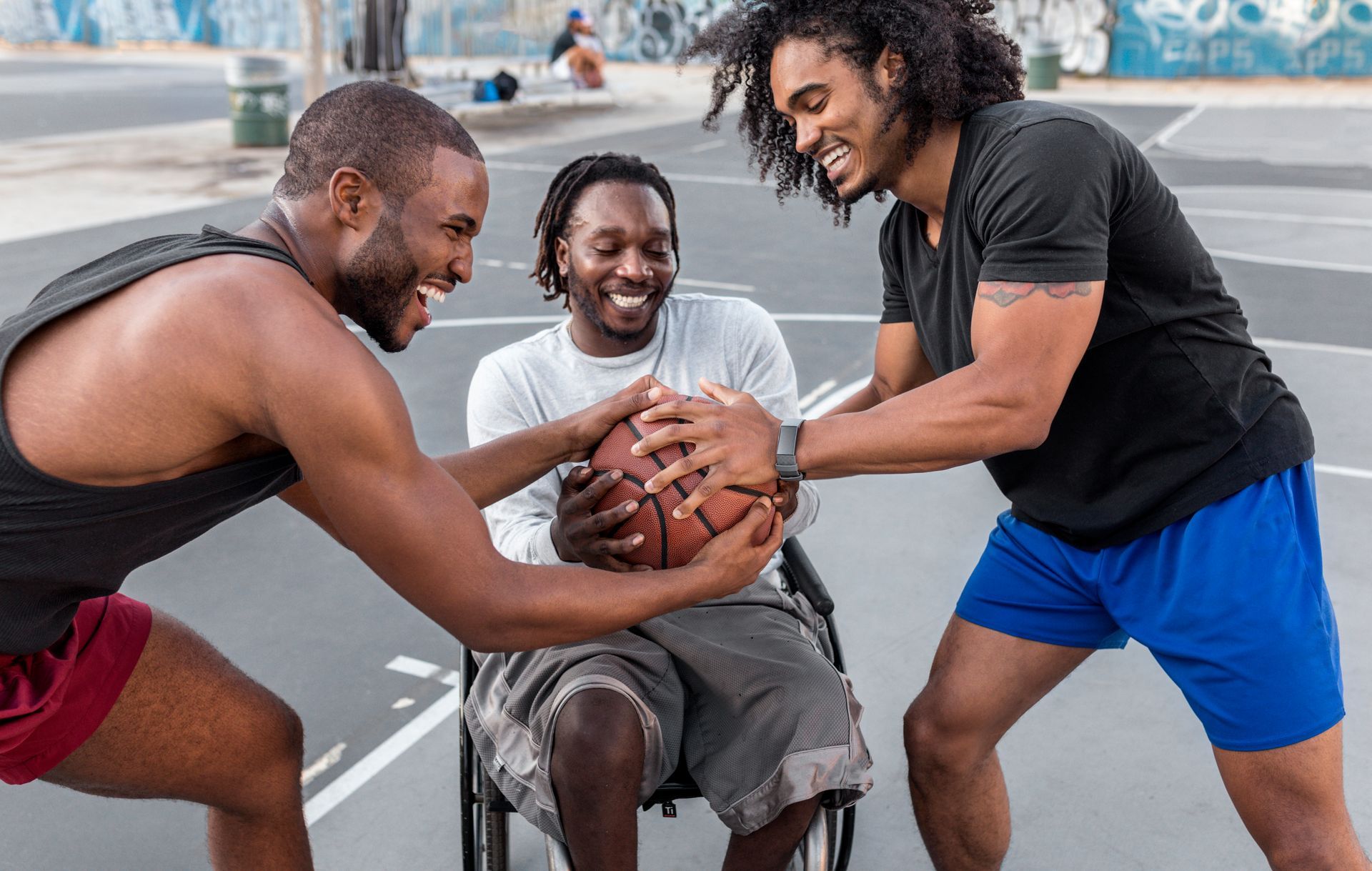 Três pessoas jogando basquete em cadeira de rodas ao ar livre, sorrindo e tentando alcançar a bola.