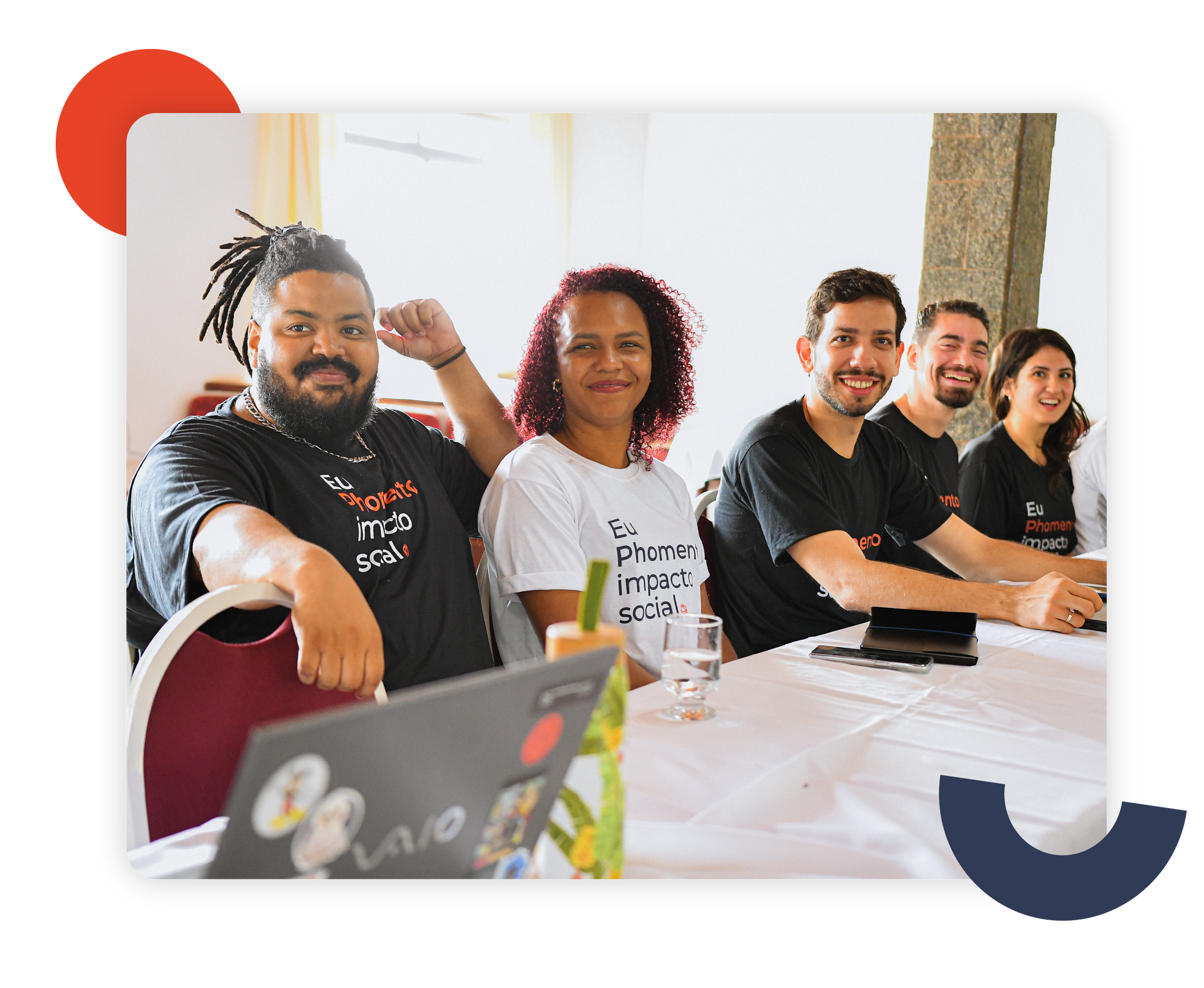 Pessoas sentadas à mesa em uma sala de reuniões iluminada, sorrindo e vestindo camisetas pretas.