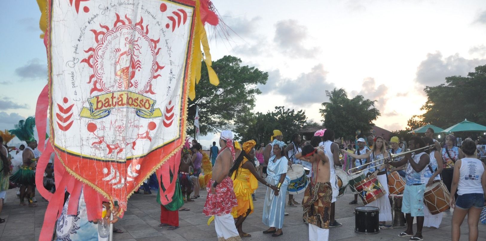 Pessoas tocando instrumentos de percussão e dançando em uma procissão cultural festiva ao ar livre, com uma faixa decorativa.