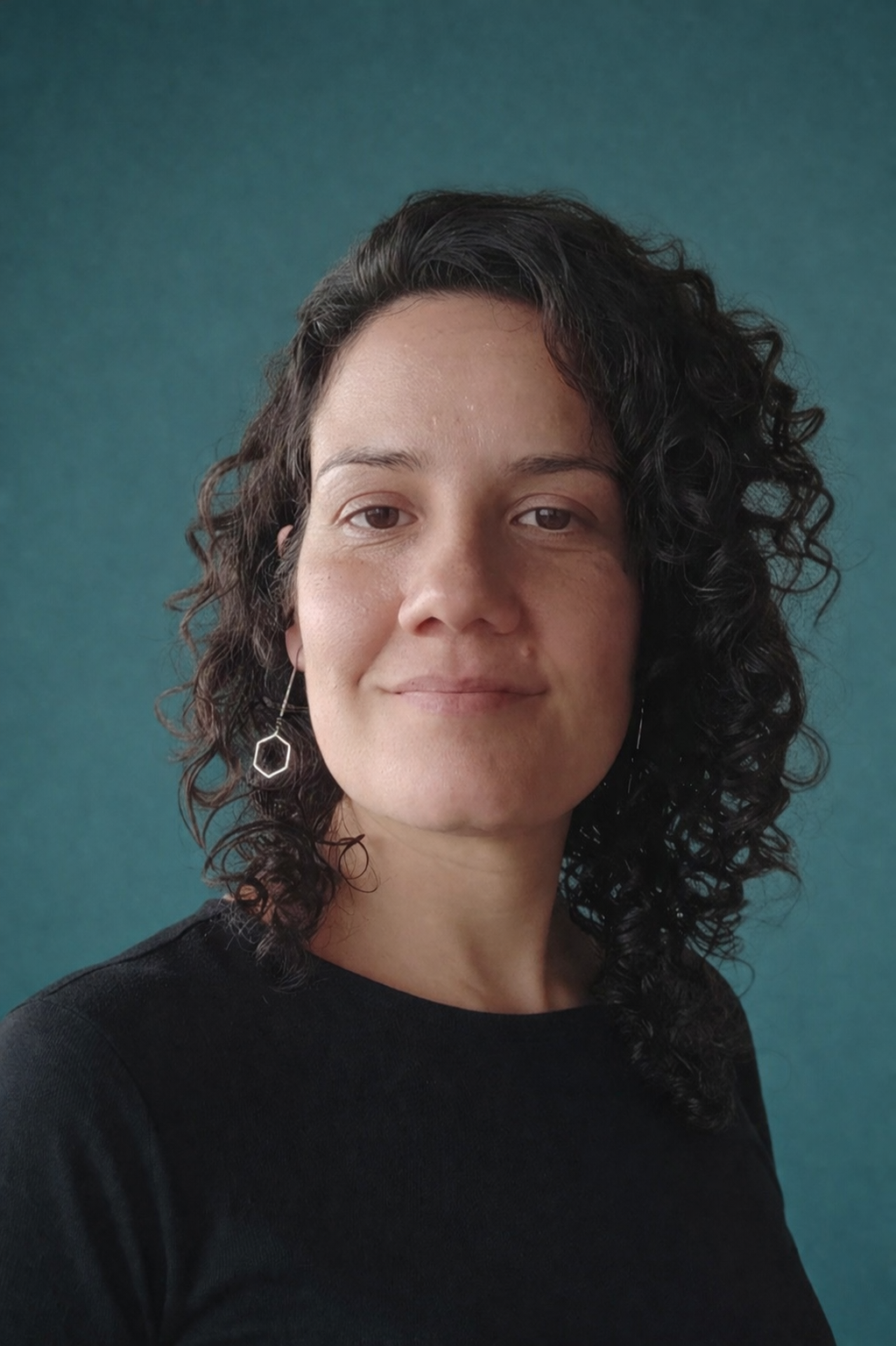 Retrato de uma pessoa com cabelo escuro encaracolado vestindo uma blusa preta contra um fundo azul-petróleo.