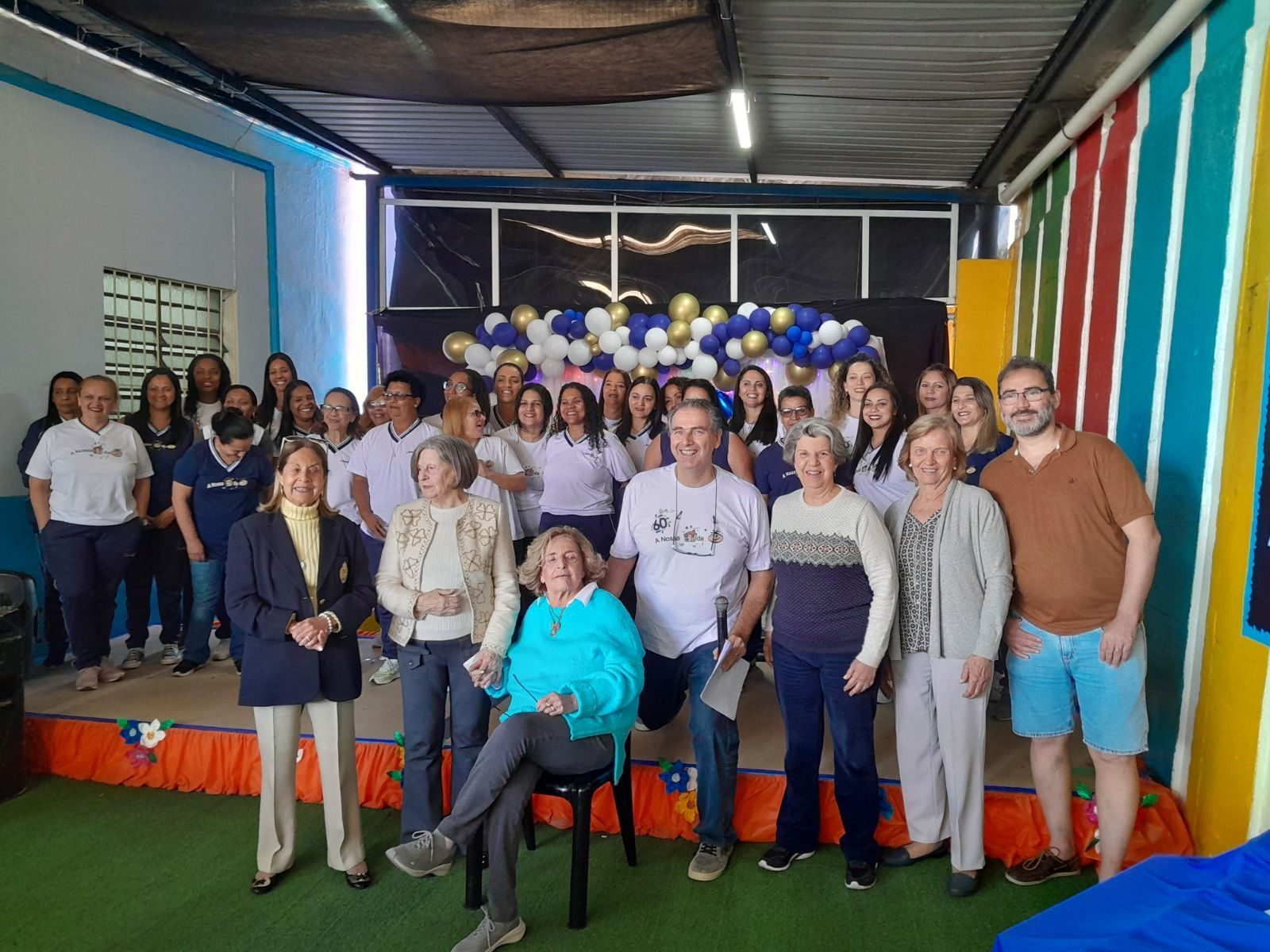 Um grupo de pessoas em pé, posando para fotos em um espaço interno decorado com arcos de balões azuis e dourados.