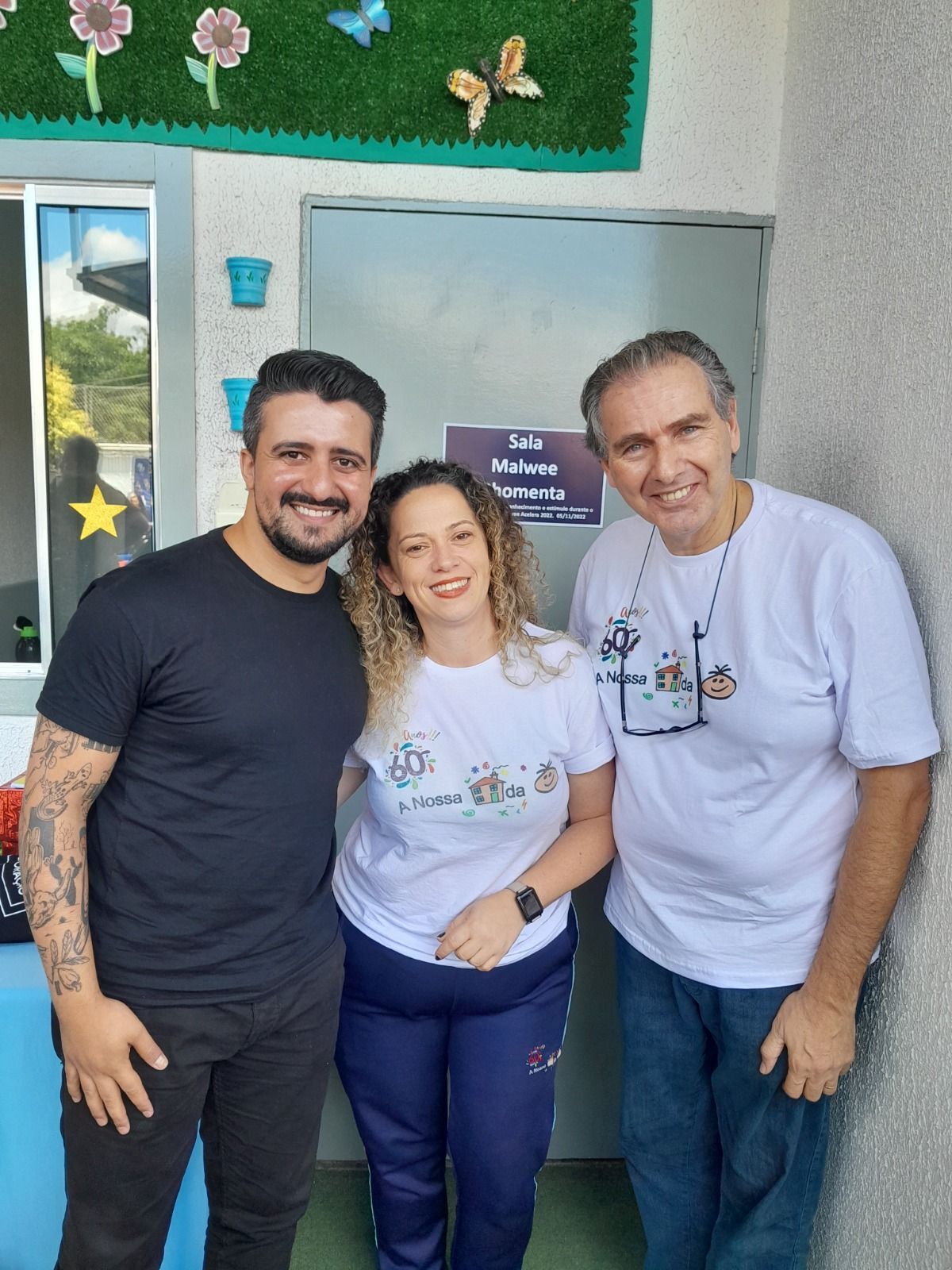 Três pessoas em pé, sorrindo, em frente a uma porta decorada com flores e borboletas.