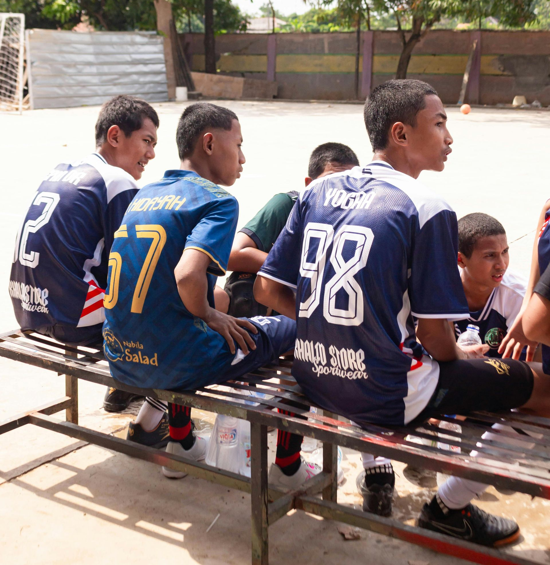 Jogadores de futebol com camisas azuis sentam-se em um banco em um campo ao ar livre, assistindo ao jogo.