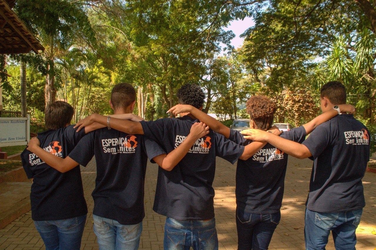 Um grupo de cinco pessoas vestindo camisas azul-escuras iguais está de pé, com os braços em volta dos ombros umas das outras, ao ar livre.
