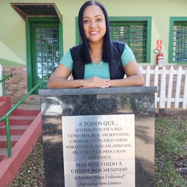 Uma pessoa sorrindo atrás de uma placa comemorativa na Cidade dos Meninos, que apresenta uma mensagem inspiradora de boas-vindas.
