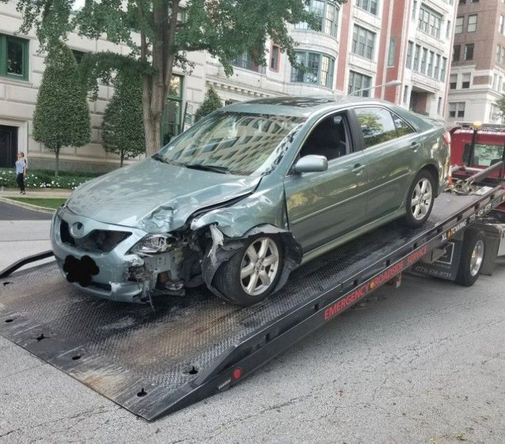 Silver car being towed on a flatbed tow truck, secured with straps.