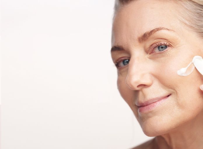 Woman applying cream to her face, smiling. White background — Aqua Marine Skin and Beauty in Tuncurry, NSW