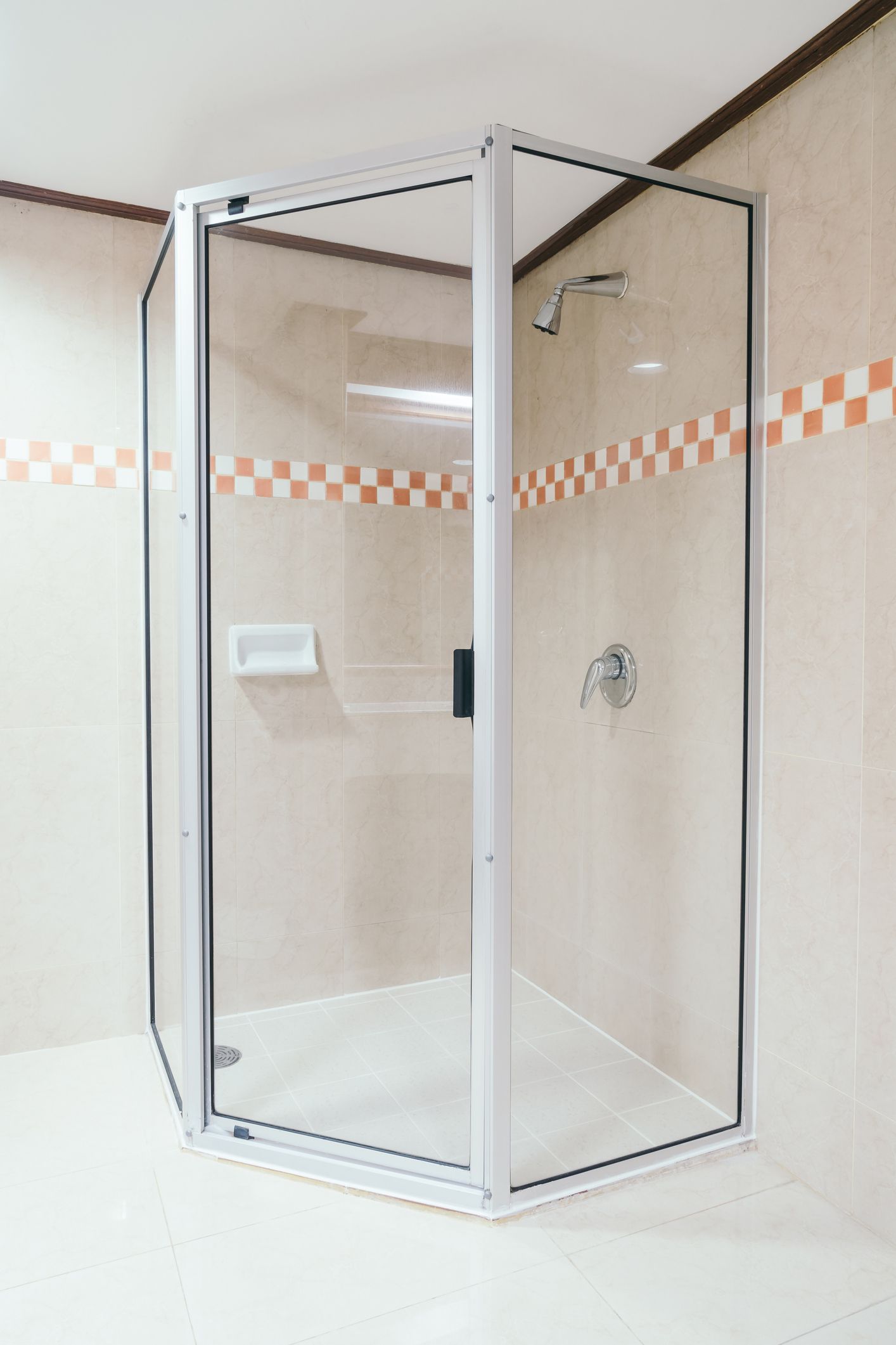 A glass corner shower enclosure with a white frame, chrome fixtures, and tan tiled walls with a checkered border.