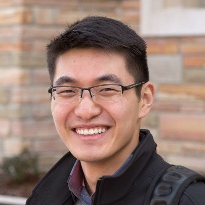 A young man wearing glasses and a black jacket is smiling.