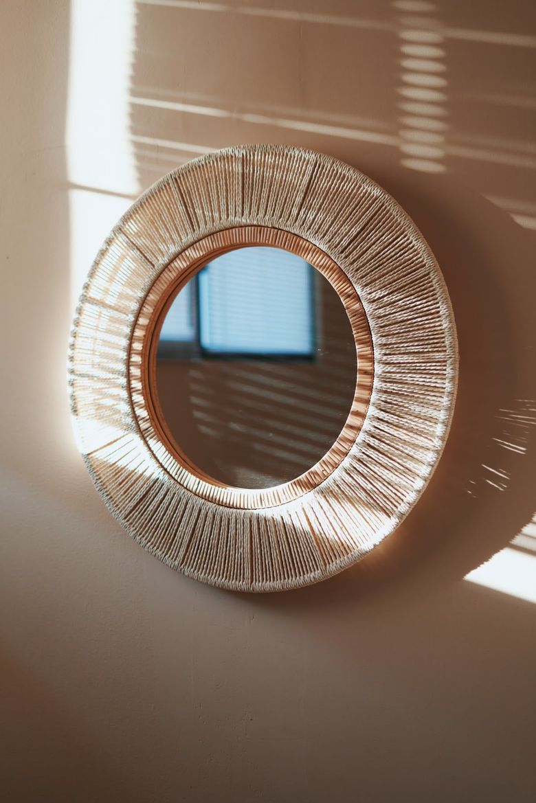 Specchio rotondo da parete intrecciato, con la luce del sole che proietta ombre a strisce su una parete beige.