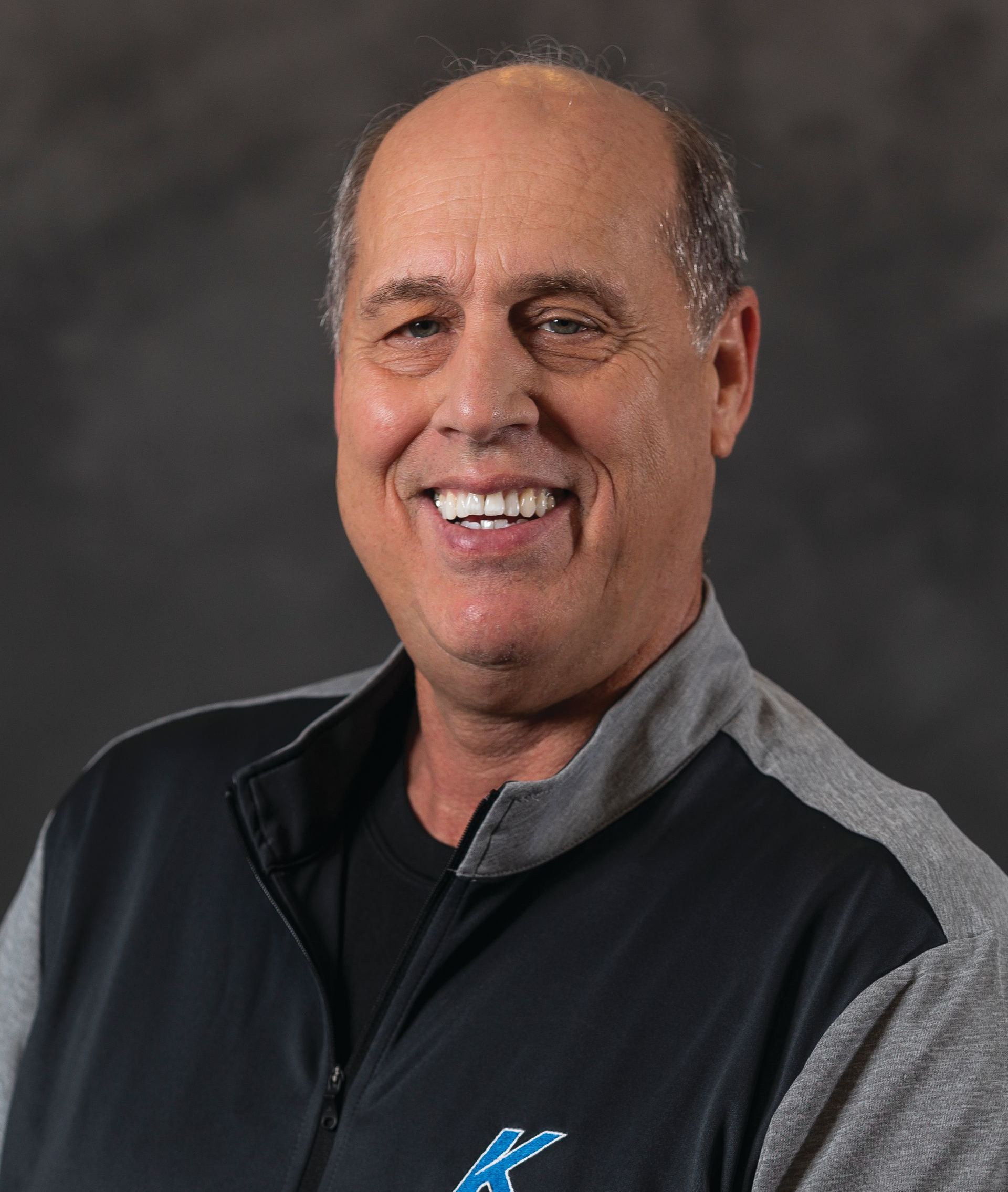 Smiling man in a black and gray jacket against a dark studio backdrop