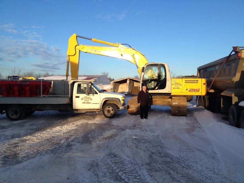 Un homme se tient à côté d’une excavatrice jaune et d’un camion dans la neige.