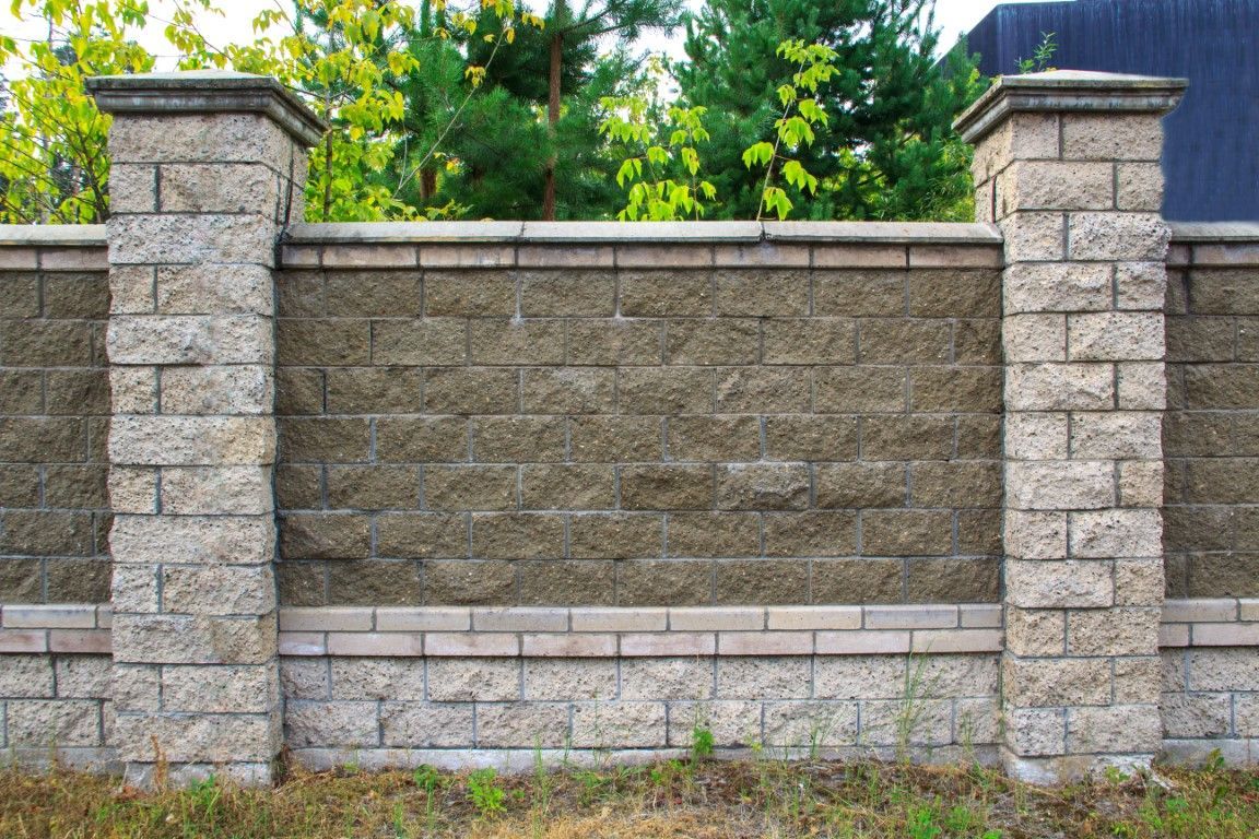 Stone wall with pillars against a backdrop of trees and a building.