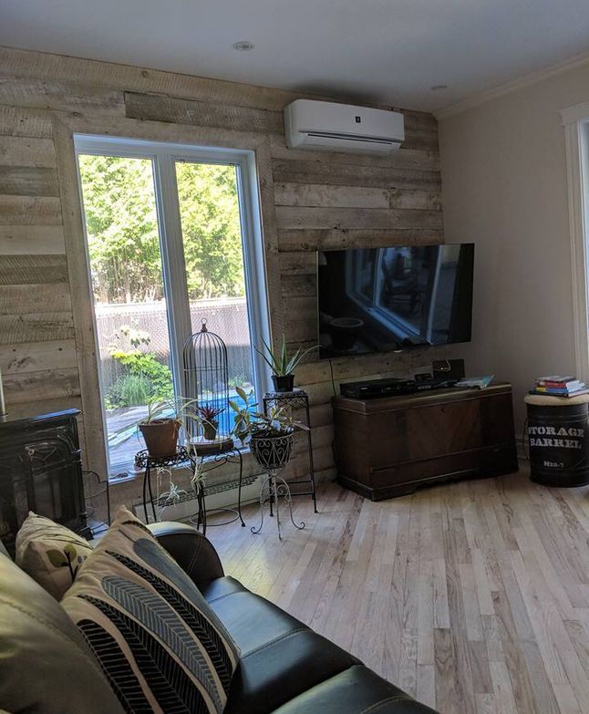 Salon confortable avec canapé noir, télévision sur meuble en bois, grande fenêtre et mur d'accent en bois rustique