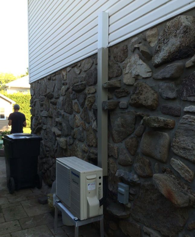Côté d'une maison aux fondations en pierre, unité de climatisation extérieure et une personne près de poubelles dans une ruelle étroite.