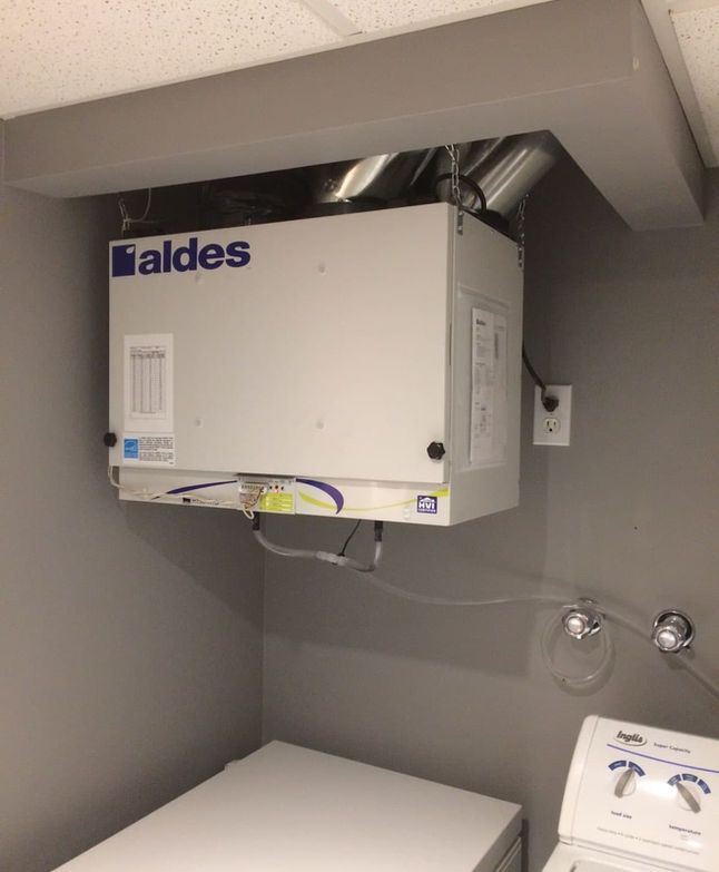 Unité de ventilation White Aldes installée dans une petite buanderie grise au-dessus d'une armoire.