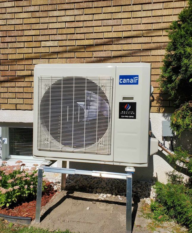 Unité de pompe à chaleur extérieure montée sur un support à côté d'une maison en briques, avec une grille de ventilation et une étiquette de marque bleue.