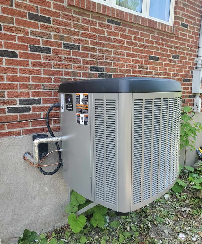 Unité de climatisation extérieure installée à côté d'un mur de maison en briques, avec tuyauterie en cuivre apparente et grilles de ventilation.