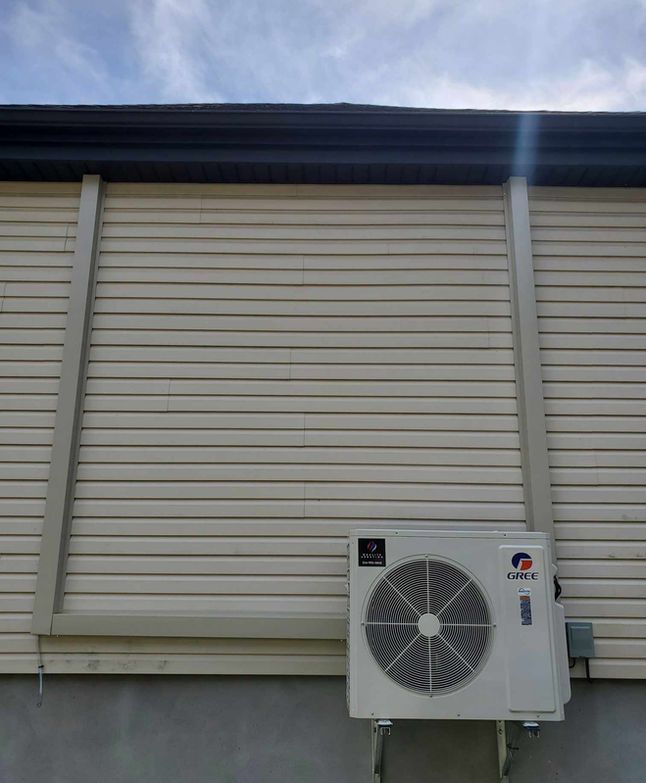 Climatiseur blanc fixé sur un mur beige d'une maison, sous un avant-toit sombre.