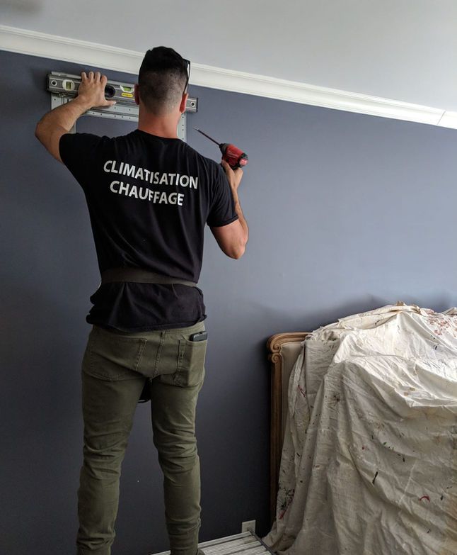 Un technicien mesure un mur gris à l'aide d'un niveau et d'un mètre ruban, à côté d'une chaise recouverte d'un tissu.