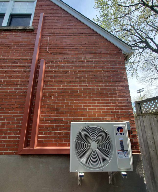 Pompe à chaleur extérieure murale sur une maison en briques rouges, à côté d'une fenêtre et de la toiture.