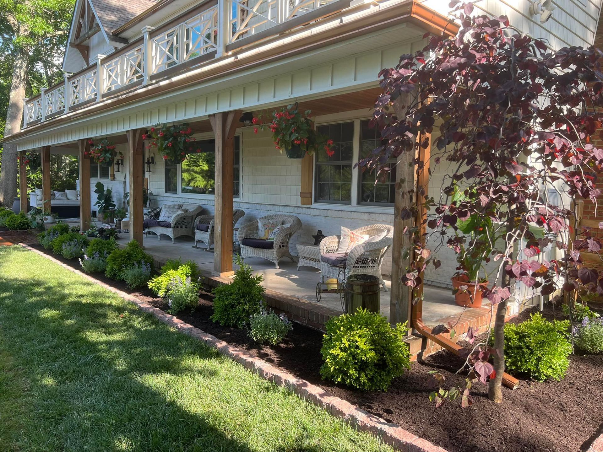Victorian house with wraparound porch and garden beds, featuring white wicker furniture and a vibrant red-leaved tree.