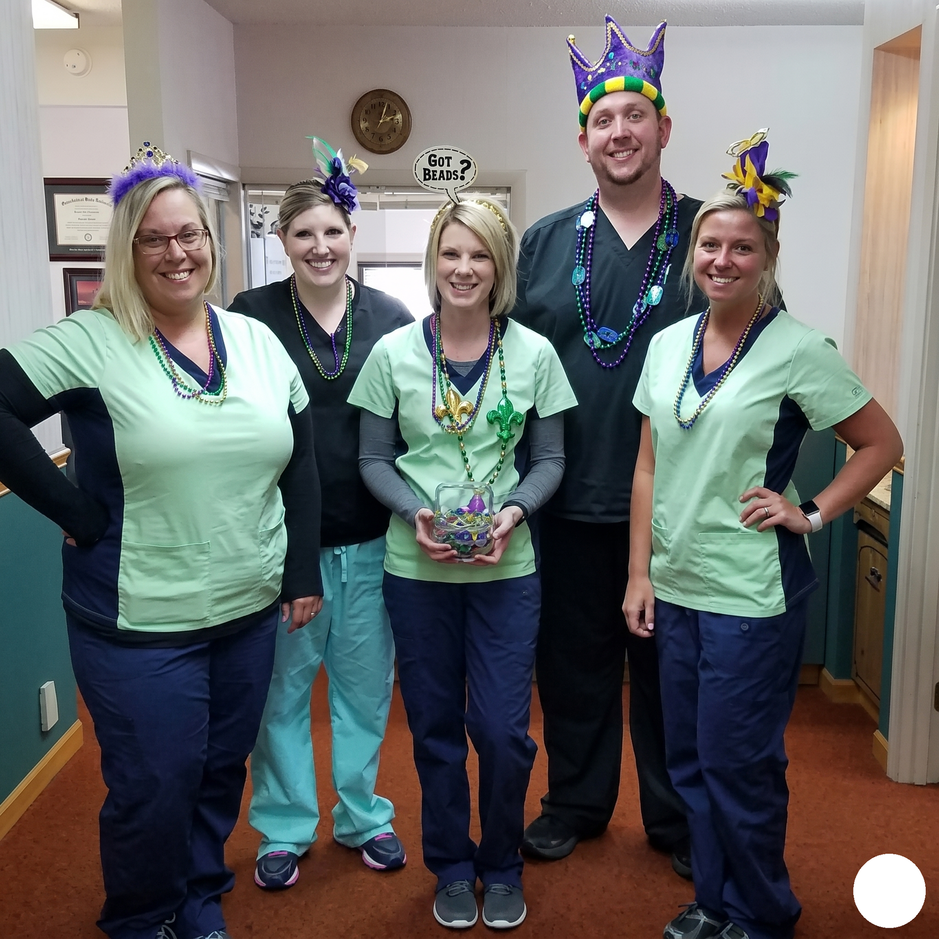 Five coworkers in festive crowns pose in a hallway, one holding a small trophy-like gift.