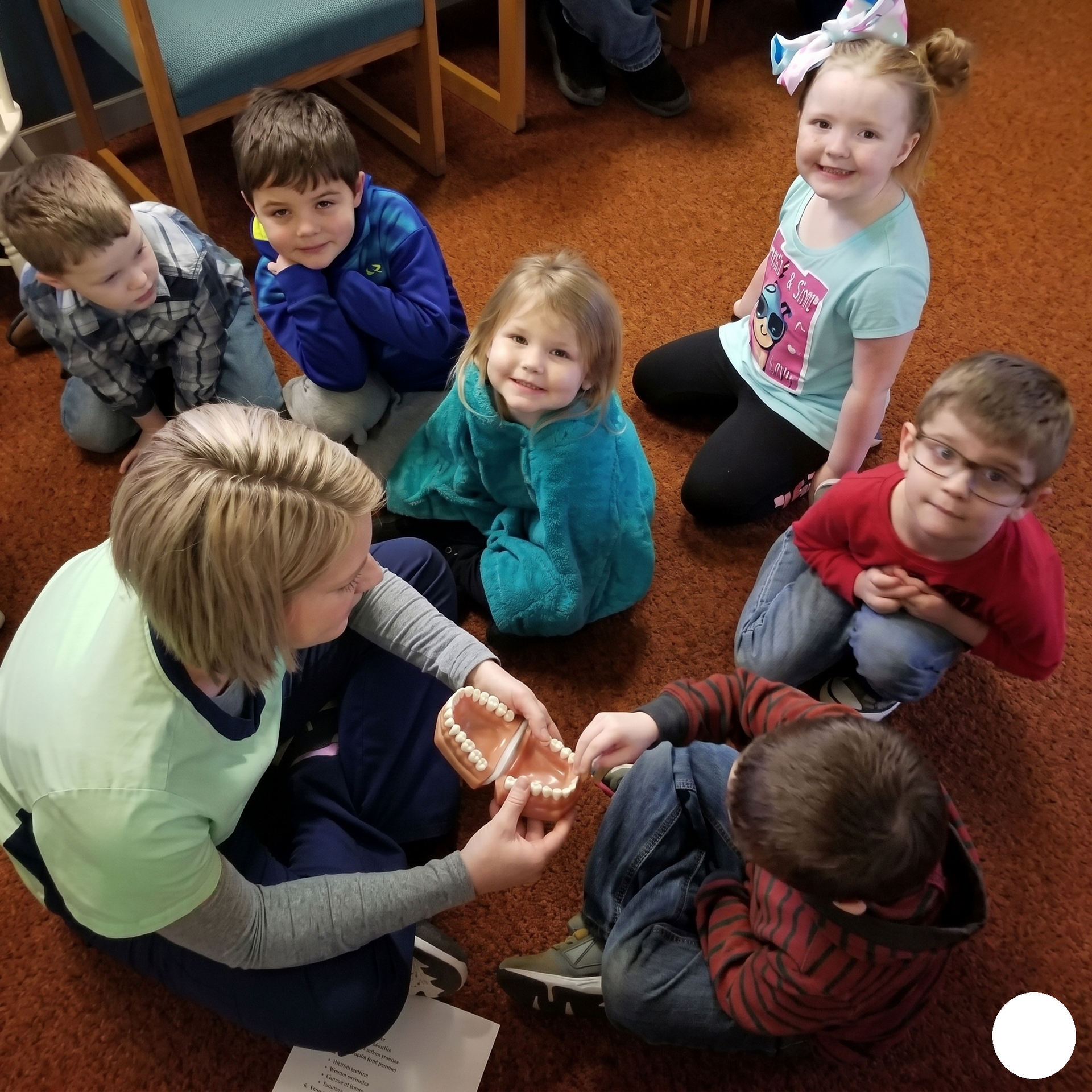 Teacher shows children a small object while they sit together on a classroom carpet.