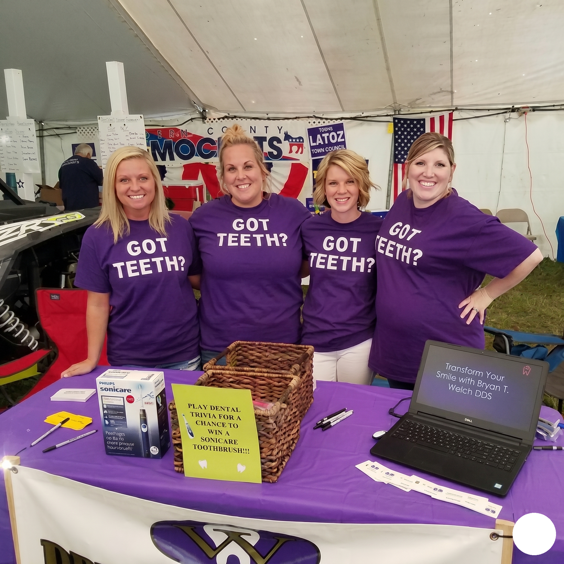 Four volunteers in purple “Got Teeth?” shirts at a booth with flyers and a laptop under a white tent.