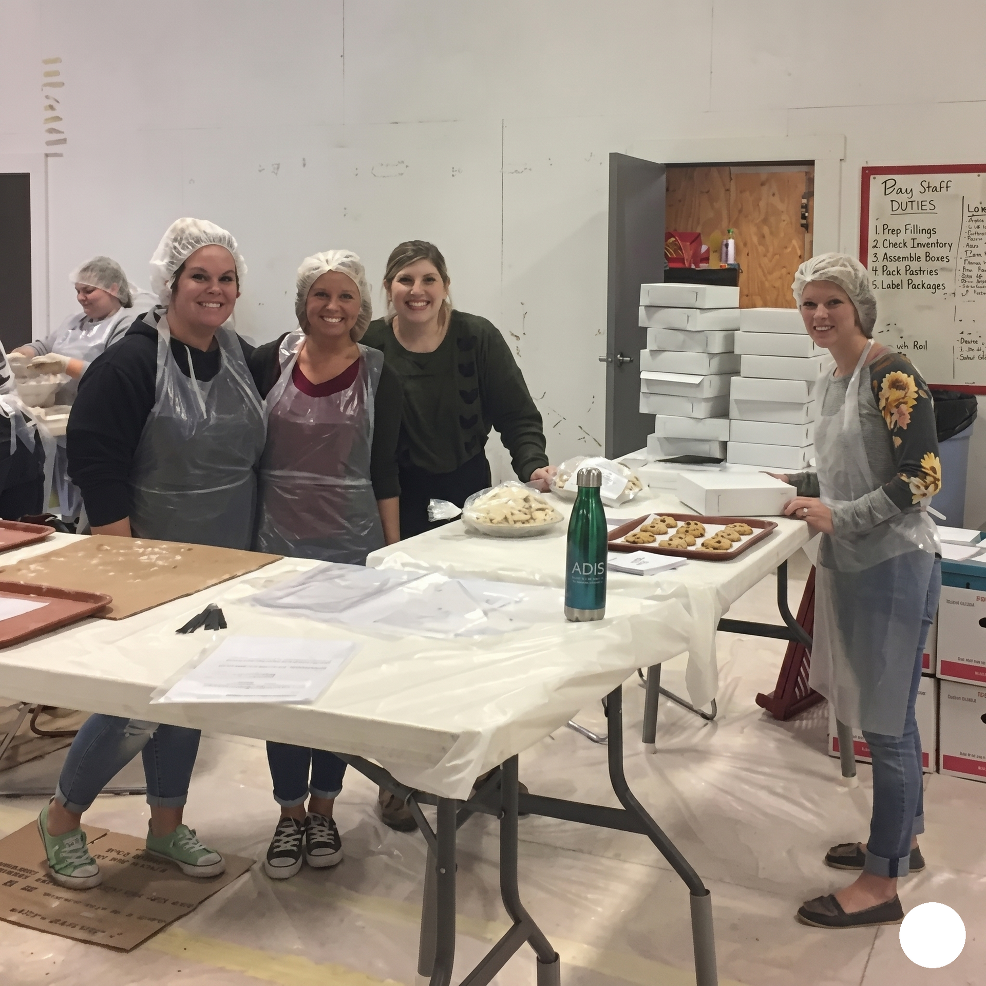 Five people in aprons smile behind a table with food trays in a bright kitchen or community room.