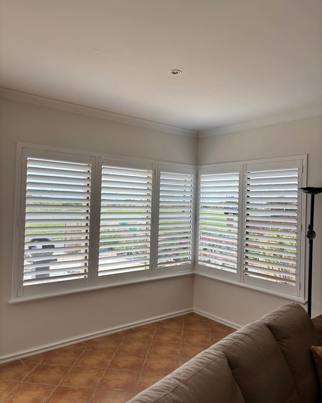 Bright corner living room with white plantation shutters, tiled floor, and part of a beige sofa.