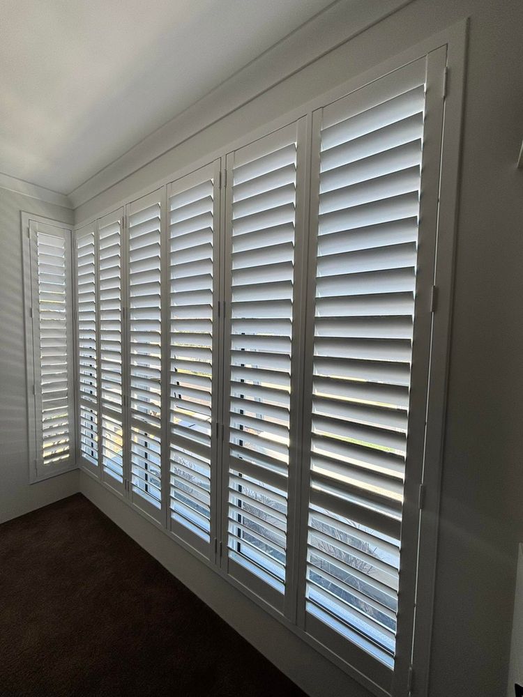 Row of white plantation shutters covering tall windows in a bright room in Perth, WA.