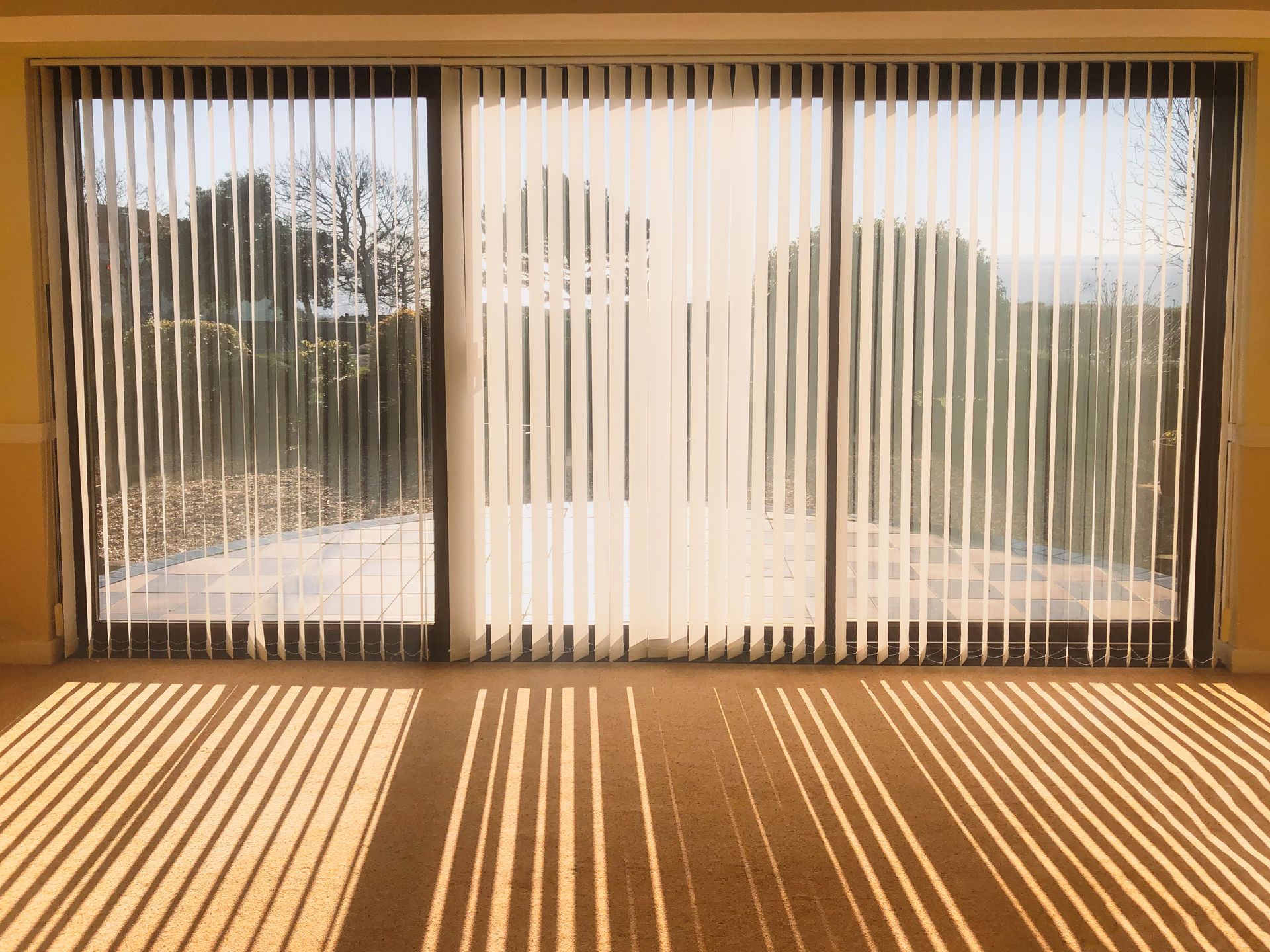Sunlit room with vertical blinds casting striped shadows across the floor, viewed through large windows