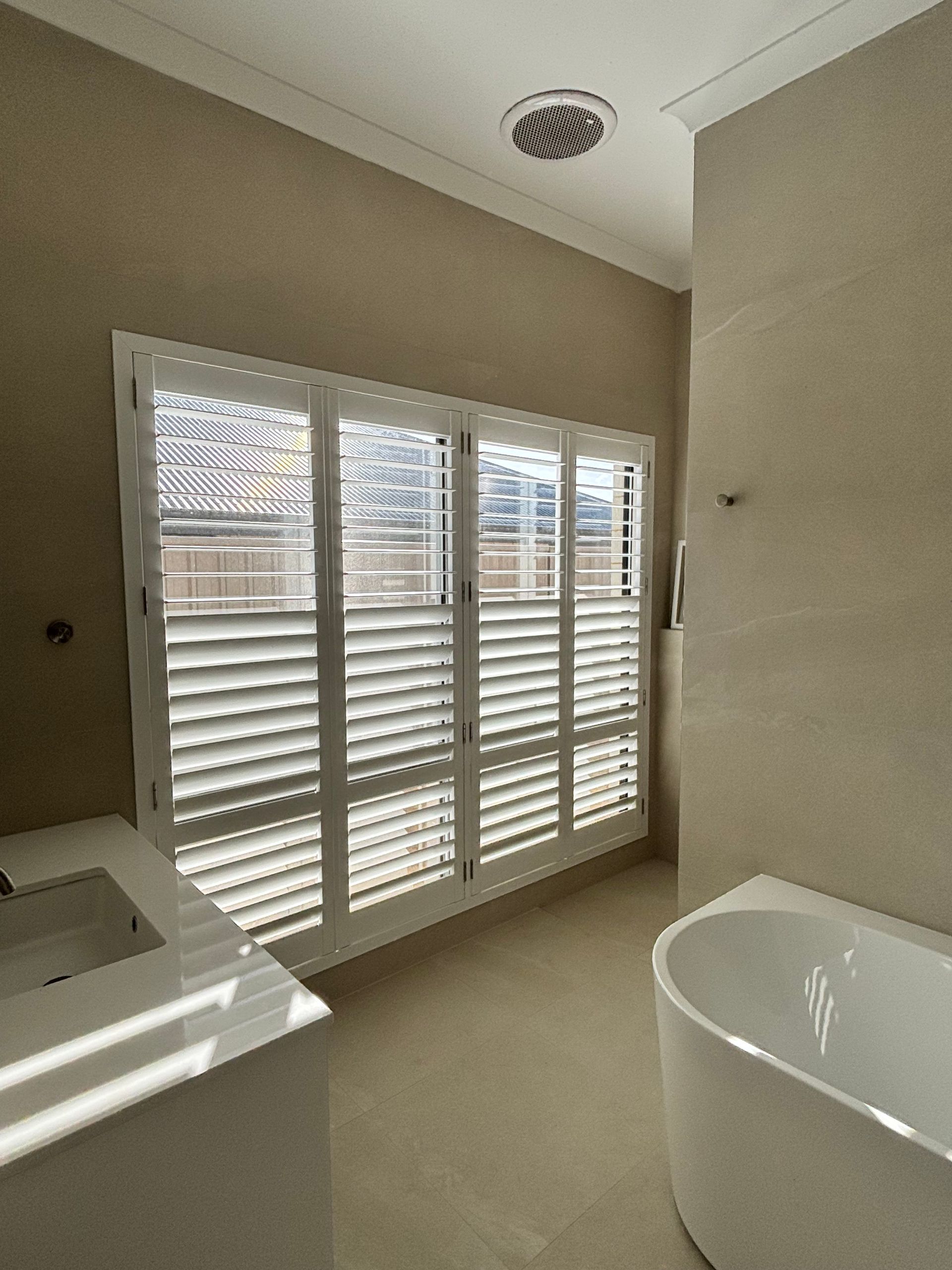 Plantation shutters installed in a bright white room with a round window, ceiling fan, and floor cushions by the wall in Joondalup, WA.