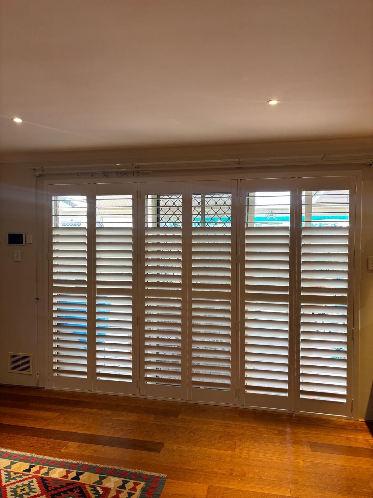 Plantation shutters installed in a bright room with white plantation shutters around a corner window and French doors, hardwood floors, and blue outside in Perth, WA.