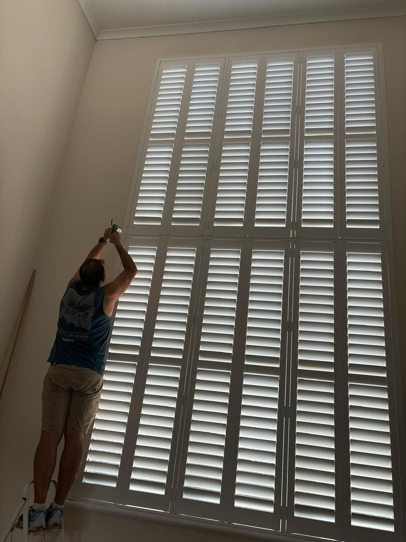 Plantation shutters installed in a person using a tool to reach tall white window shutters in a bright room in Joondalup, WA.