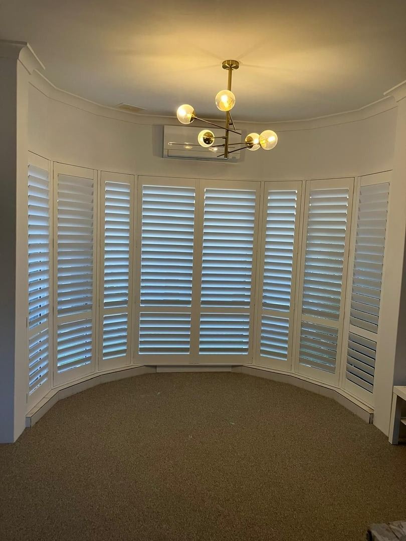 Plantation shutters installed in a curved bay window with white shutters, beige carpet, and a brass ceiling light fixture in Joondalup, WA.