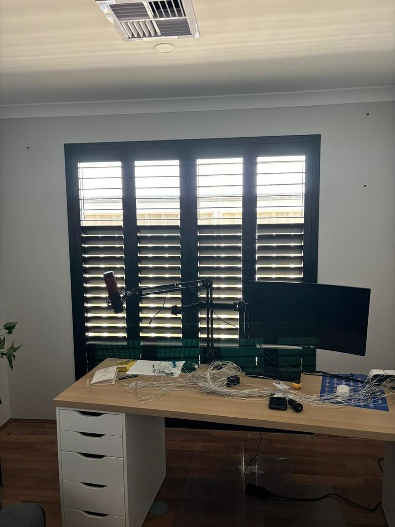 Plantation shutters installed in a home office with desk, black shutters, computer monitor, and office supplies by a window in Perth, WA.
