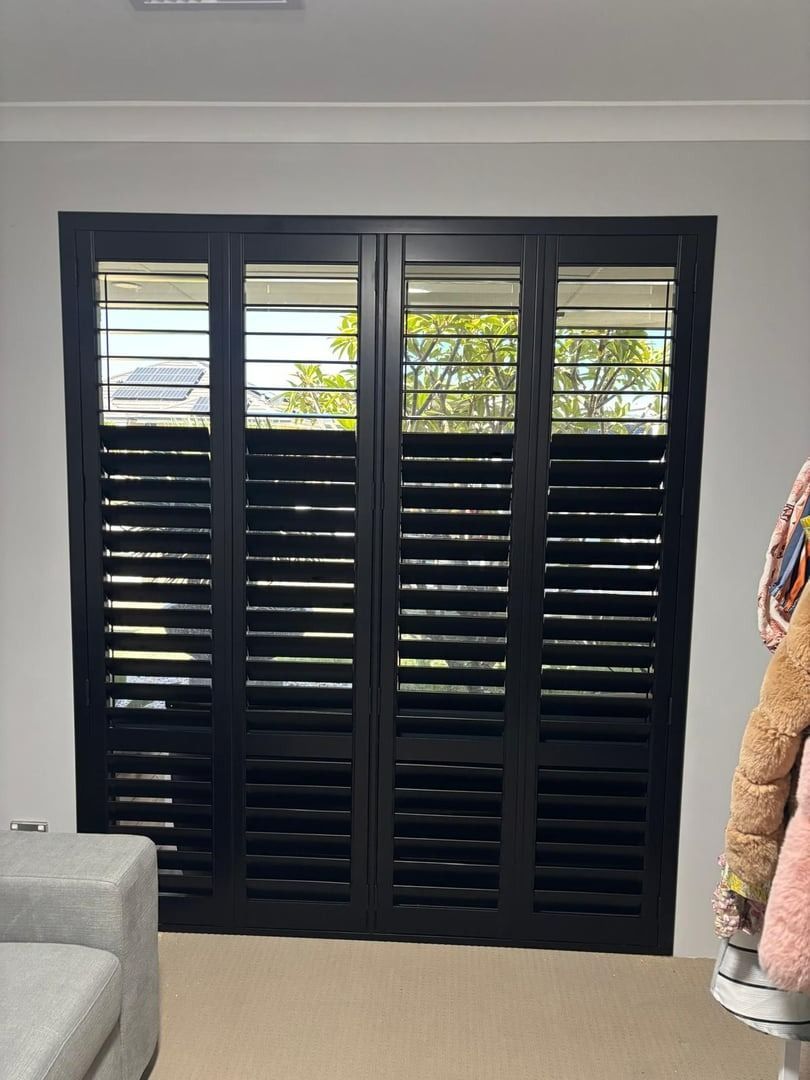 Plantation shutters installed in a black louvered window shutters in a white room, with a couch edge and coat rack visible in Perth, WA.