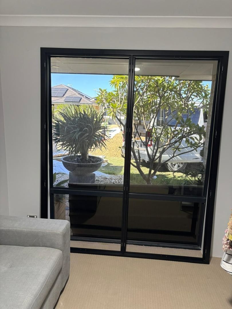 Plantation shutters installed in a living room with large black sliding glass doors opening to a sunny patio and greenery in Joondalup, WA.