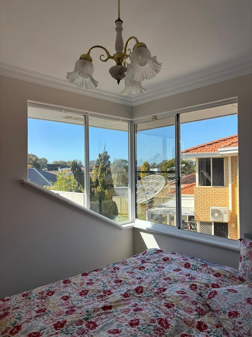 Plantation shutters installed in a bright bedroom with bay windows, floral bedspread, chandelier, and a view of neighboring houses in Joondalup, WA.