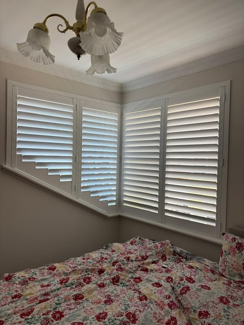 Plantation shutters installed in a bedroom corner with white shutters, beige walls, floral bedspread, and a ceiling light fixture in Joondalup, WA.