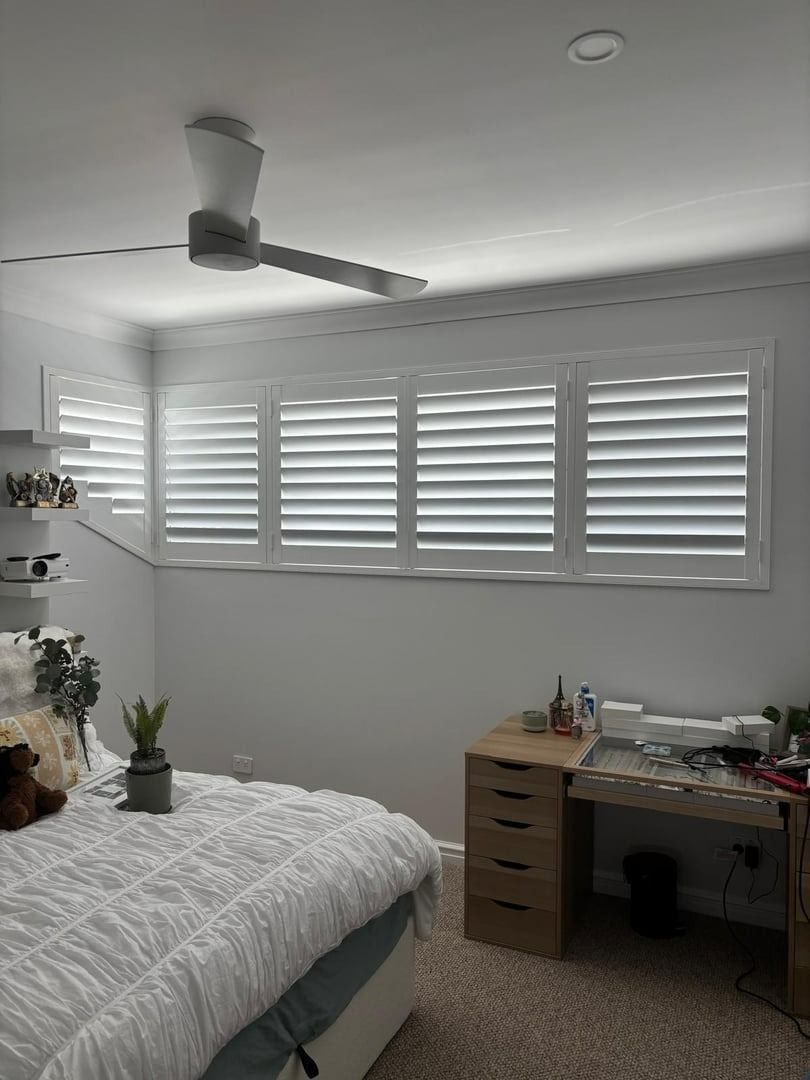 Plantation shutters installed in a bright bedroom with a bed, ceiling fan, desk, and wide windows with white shutters in Perth, WA.