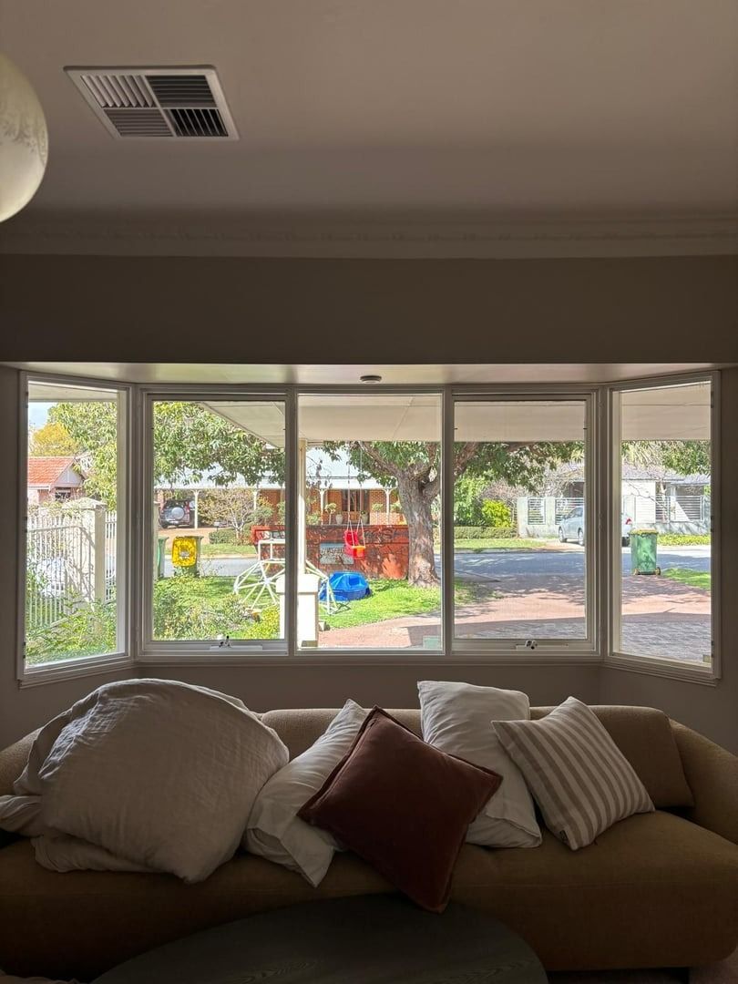 Plantation shutters installed in a living room couch facing bright bay windows with a colorful backyard view in Perth, WA.