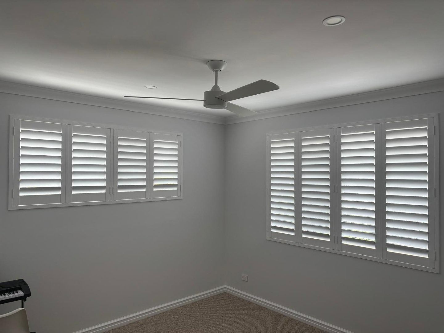 Plantation shutters installed in an empty gray room with two windows, white shutters, and a ceiling fan under recessed lights in Joondalup, WA.