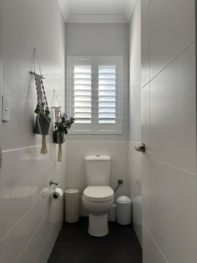 Plantation shutters installed in a narrow white bathroom with toilet, window shutters, and hanging towels and plants in Perth, WA.