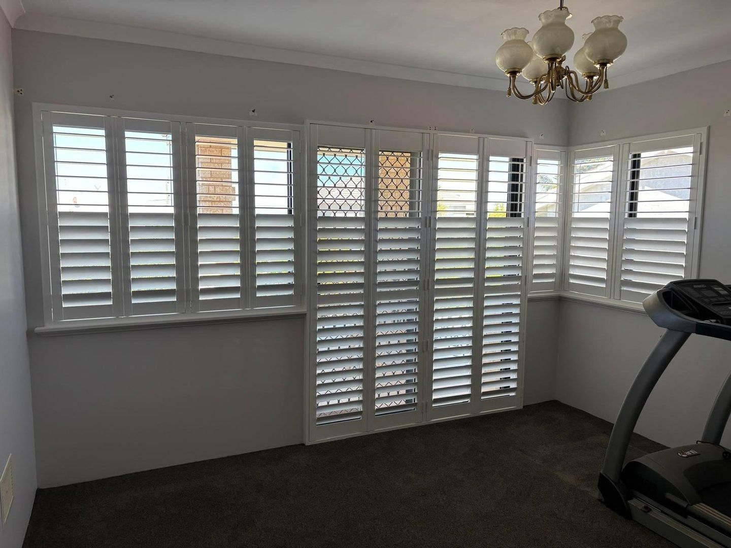 Plantation shutters installed in a bright room with white plantation shutters, chandelier, and a treadmill on dark carpet in Joondalup, WA.