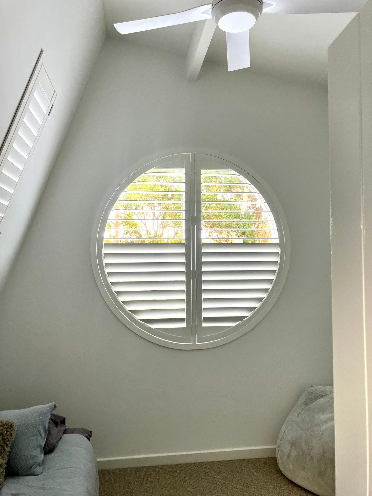 Plantation shutters installed in a bright white room with a round window, ceiling fan, and floor cushions by the wall in Perth, WA.