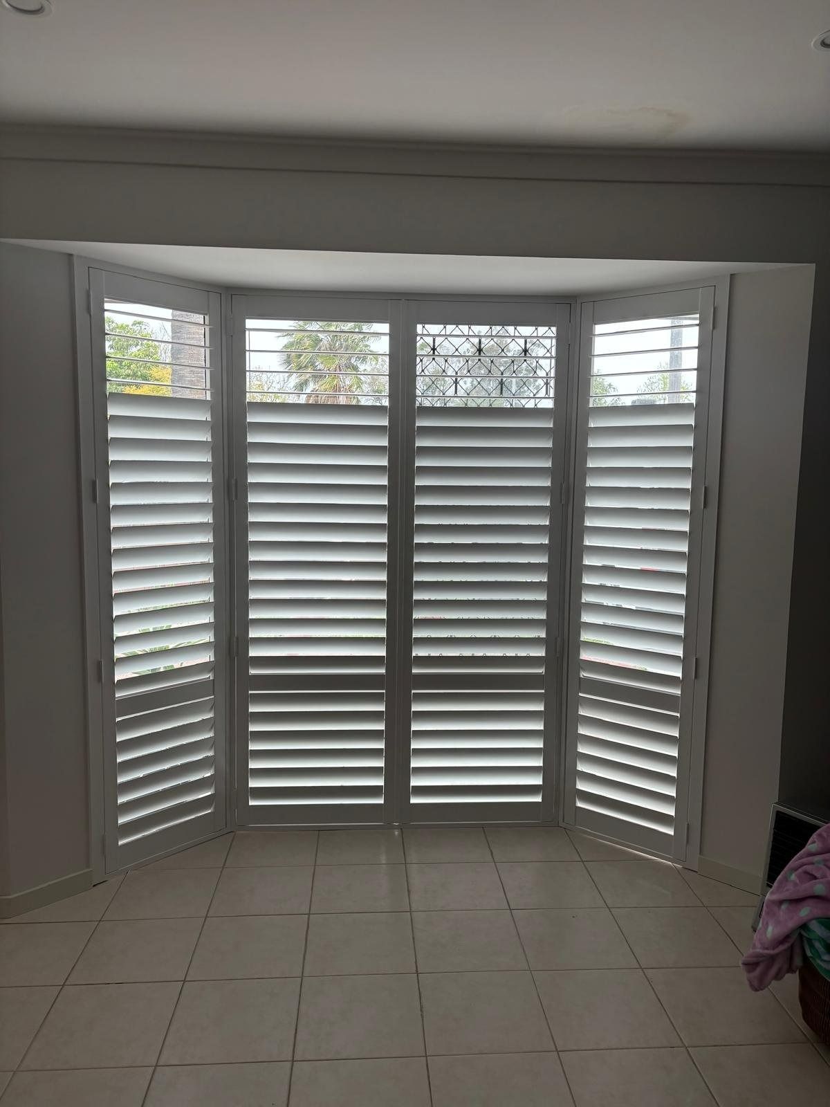 Plantation shutters installed in a living room with white tiled floor and a bay window fitted with white louvered shutters in Joondalup, WA.